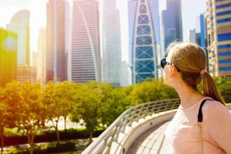 Traveler wearing sunglasses looks at Abu Dhabi city skyline from a curved pedestrian bridge on a sunny day.